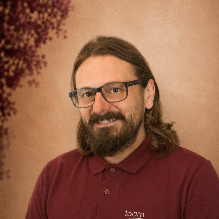 Mann mit Brille und langen Haaren, grinsend vor einem Naturhintergrund.
