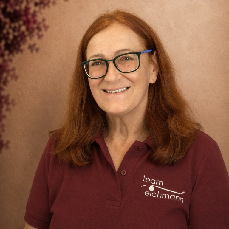 Frau mit langen roten Haaren und Brille, lächelnd, in einer natürlichen Umgebung.